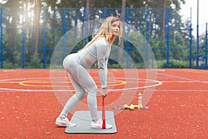 Young blond beautiful slim fit girl in gray sportswear doing squats and stretching exercise with red fitness training