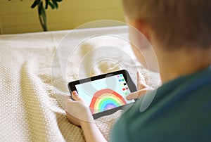 A young blond boy lying on a couch drew a rainbow on a tablet computer in his room. Education and play
