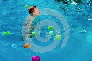 Young blond boy playing in a pool with water balloons, colorful, in summer