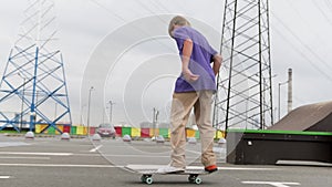 Young blond boy skateboarder rides forward on skateboard in skate park in cloudy weather. Professional skater teen