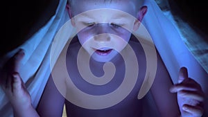 Young blond boy under covers playing on tablet a computer game.