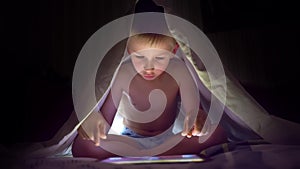 Young blond boy under covers playing on tablet a computer game.