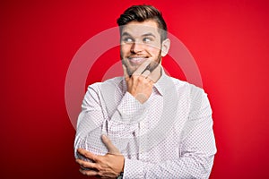 Young blond businessman with beard and blue eyes wearing elegant shirt over red background with hand on chin thinking about