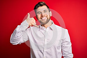 Young blond businessman with beard and blue eyes wearing elegant shirt over red background smiling doing phone gesture with hand