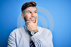 Young blond businessman with beard and blue eyes wearing elegant shirt and tie standing with hand on chin thinking about question,