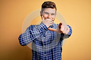 Young blond businessman with beard and blue eyes wearing shirt over yellow background laughing at you, pointing finger to the