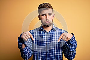 Young blond businessman with beard and blue eyes wearing shirt over yellow background Pointing down looking sad and upset,