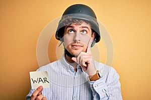 Young blond businessman with curly hair wearing helmet holding paper with war message serious face thinking about question, very