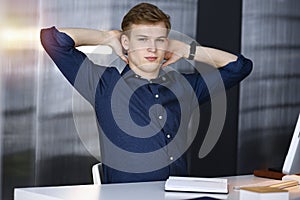 Young blond businessman and programmer stretching hands after working hard with computer in a darkened office, glare of