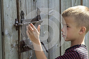 Young blond cute boy trying to open rusty slide bolt lock on lit by sun closed old wooden barn door. Children curiosity, love for