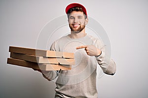 Young blond delivery man with beard and blue eyes holding cardboards of pizza very happy pointing with hand and finger