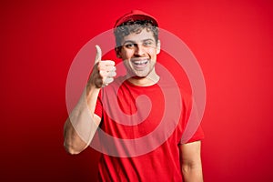 Young blond delivery man with curly hair wearing cap standing over isolated red background doing happy thumbs up gesture with hand