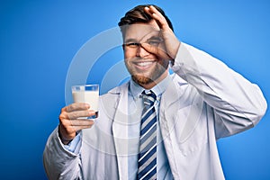 Young blond doctor man with beard and blue eyes wearing coat drinking glass of milk with happy face smiling doing ok sign with
