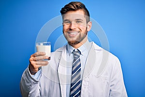 Young blond doctor man with beard and blue eyes wearing coat drinking glass of milk with a happy face standing and smiling with a