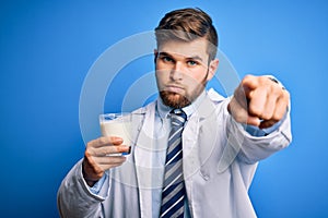 Young blond doctor man with beard and blue eyes wearing coat drinking glass of milk pointing with finger to the camera and to you,