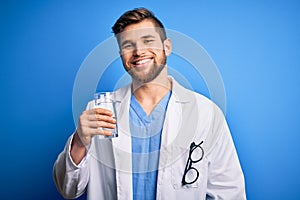 Young blond doctor man with beard and blue eyes wearing coat drinking glass of water with a happy face standing and smiling with a