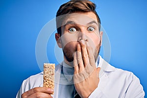 Young blond doctor man with beard and blue eyes wearing coat eating granola bar cover mouth with hand shocked with shame for