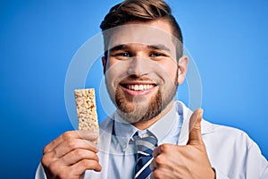 Young blond doctor man with beard and blue eyes wearing coat eating granola bar happy with big smile doing ok sign, thumb up with