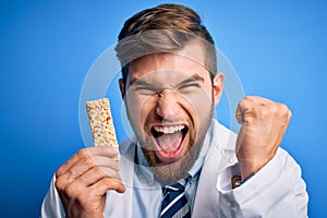 Young blond doctor man with beard and blue eyes wearing coat eating granola bar screaming proud and celebrating victory and