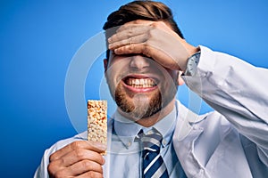 Young blond doctor man with beard and blue eyes wearing coat eating granola bar stressed with hand on head, shocked with shame and