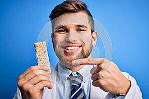 Young blond doctor man with beard and blue eyes wearing coat eating granola bar very happy pointing with hand and finger