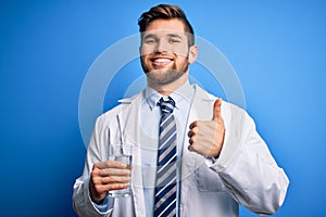 Young blond doctor man with beard and blue eyes wearing coat and tie drinking glass of water happy with big smile doing ok sign,