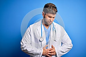 Young blond doctor man with beard and blue eyes wearing white coat and stethoscope with hand on stomach because indigestion,