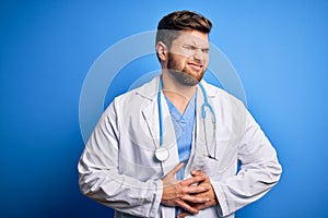 Young blond doctor man with beard and blue eyes wearing white coat and stethoscope with hand on stomach because nausea, painful