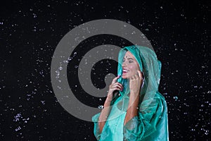 Young Blond Female in Green Raincoat Posing Under The Water Drops Against Black Background