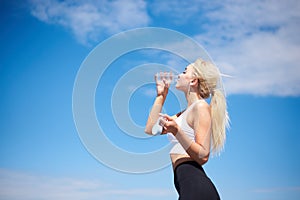 Young blond fit woman, wearing black leggings and white top,drinking water from glass bottle outside with blue sunny sky on