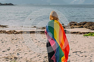 Young blond girl covered with LGBTQI flag  walking in the day on the white beach near the sea