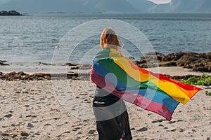 Young blond girl covered with LGBTQI flag  walking in the day on the white beach near the sea