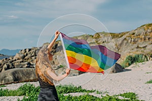 Young blond girl holding  LGBTQI flag  walking in the day on the white beach near the sea