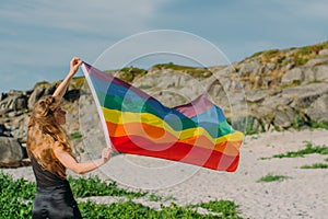 Young blond girl holding  LGBTQI flag  walking in the day on the white beach near the sea