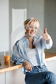 A young blond girl in stylish glasses sits in the kitchen with a glass of juice. Looks at the camera and shows the super gesture