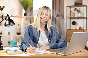 Young blond girl wearing headset making notes and smiling, sitting at the desk at bright room