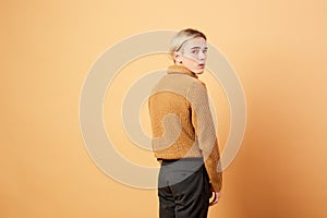 Young blond guy dressed in mustard color sweater and black pants is posing in the studio on the beige background