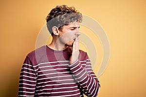 Young blond handsome man with curly hair wearing casual striped sweater bored yawning tired covering mouth with hand