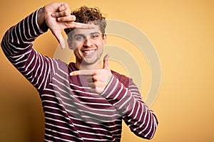 Young blond handsome man with curly hair wearing casual striped sweater smiling making frame with hands and fingers with happy