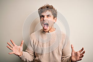 Young blond handsome man with curly hair wearing casual sweater over white background crazy and mad shouting and yelling with