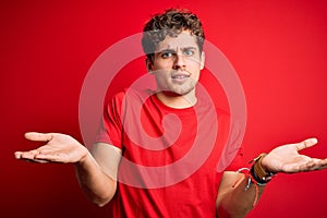 Young blond handsome man with curly hair wearing casual t-shirt over red background clueless and confused with open arms, no idea