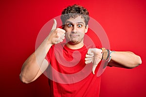 Young blond handsome man with curly hair wearing casual t-shirt over red background Doing thumbs up and down, disagreement and