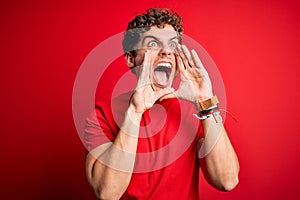 Young blond handsome man with curly hair wearing casual t-shirt over red background Shouting angry out loud with hands over mouth