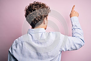 Young blond handsome man with curly hair wearing striped shirt over white background Posing backwards pointing ahead with finger