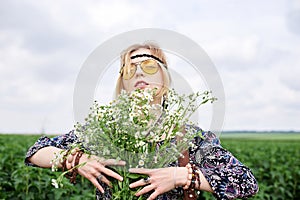 Young blond hippie woman, wearing grey boho style dress and yellow sunglasses, standing on green field, holding camomile bouquet,