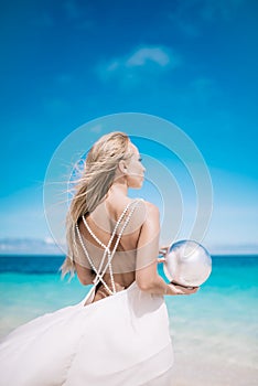 Young blond long hair bride wear a white open back wedding dress and stand on the white sand beach with a pearl. Looking to the se