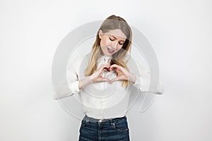 Young blond lovely woman making heart symbol with her hands standing on isolated white background, looking gentle,body language,