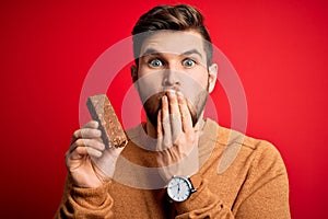 Young blond man with beard and blue eyes eating healthy chocolate protein bar cover mouth with hand shocked with shame for