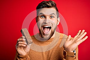 Young blond man with beard and blue eyes eating healthy chocolate protein bar very happy and excited, winner expression