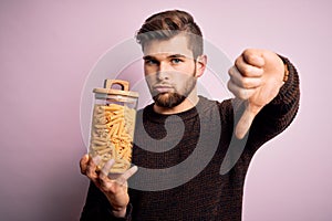 Young blond man with beard and blue eyes holding bottle of Italian dry pasta macaroni with angry face, negative sign showing